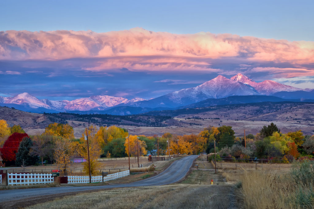 Stunning Colorado landscape backdropped by purple glowing, snowcapped mountains lit with the first rays of dawn while peering over a colorful, autumn display of trees surrounding a homestead.